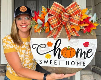 May include: A round wooden sign with the words "Home Sweet Home" and a pumpkin illustration. The sign is decorated with fall leaves and a plaid bow. The sign is approximately 12 inches in diameter.
