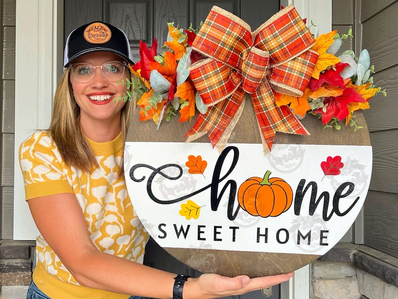 May include: A round wooden sign with a white background and black lettering that reads 'home sweet home'. The sign is decorated with a plaid bow, fall leaves, and a small pumpkin. The sign is being held by a person.