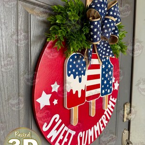 Op de afbeelding: Een rond rood houten deurbord met drie verhoogde 3D-ijslolly-uitsparingen in Amerikaanse vlagpatronen. Het bord bevat witte sterren en de witte tekst SWEET SUMMERTIME. Bovenop zit een blauwe strik met witte sterren en wat groen blad.
