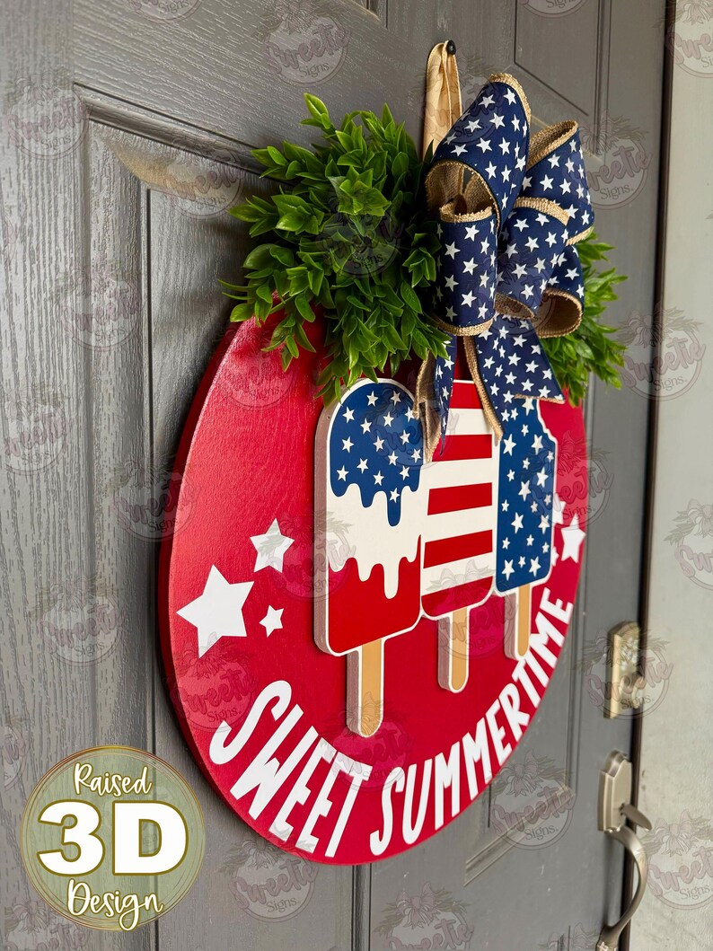 Op de afbeelding: Een rond rood houten deurbord met drie verhoogde 3D-ijslolly-uitsparingen in Amerikaanse vlagpatronen. Het bord bevat witte sterren en de witte tekst SWEET SUMMERTIME. Bovenop zit een blauwe strik met witte sterren en wat groen blad.