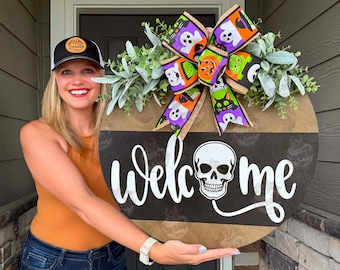 May include: A round wooden door sign featuring the text "welcome" with a white skull replacing the letter "o". The sign is decorated with a large multi-colored bow displaying ghosts, pumpkins, and cats, accented by green foliage.
