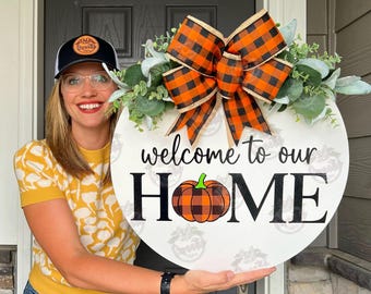 May include: A white round wooden door hanger with the text "welcome to our HOME" in black lettering. A black and orange plaid bow with green leaves is attached to the top of the door hanger. A small orange pumpkin with a black and orange plaid design is centered below the text.