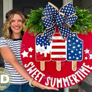 Op de afbeelding: Een rond rood houten deurbord met drie ijsjesafbeeldingen in Amerikaanse vlagpatronen, groen kunstblad en een grote blauw-witte strik van jute met sterrenpatroon. De tekst SWEET SUMMERTIME staat in witte letters aan de onderkant.