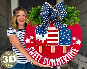 May include: A round red wooden door sign featuring three popsicle graphics with American flag patterns, green faux foliage, and a large blue and white star-patterned burlap bow. The text SWEET SUMMERTIME is printed in white at the bottom.