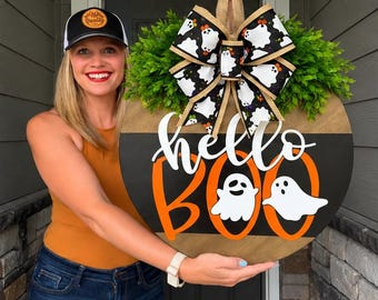 May include: A round wooden door sign featuring the text hello BOO in white and orange lettering with two white ghost illustrations. The sign is decorated with a large bow featuring a ghost pattern and green faux foliage.