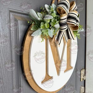 Puede incluir: Un letrero de puerta de madera con una gran letra "M" blanca sobre un fondo marr&oacute;n. El letrero est&aacute; decorado con vegetaci&oacute;n y un lazo a rayas blanco y negro. Se adjunta una cuerda de yute para colgarlo.