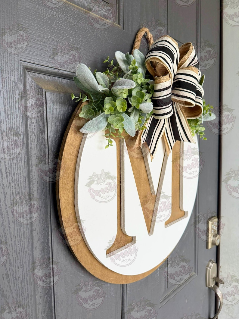 Puede incluir: Un letrero de puerta de madera con una gran letra "M" blanca sobre un fondo marr&oacute;n. El letrero est&aacute; decorado con vegetaci&oacute;n y un lazo a rayas blanco y negro. Se adjunta una cuerda de yute para colgarlo.