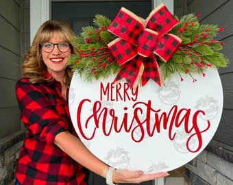May include: A round white Christmas sign with the words "Merry Christmas" in red script. The sign is decorated with a red and black plaid bow, greenery, and red berries. The sign is held by a person wearing a red and black plaid shirt.