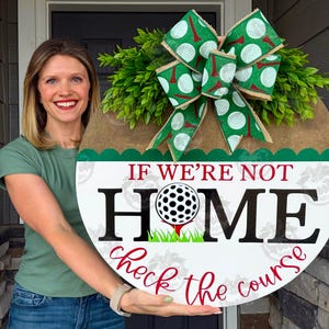 May include: Round wooden golf-themed door sign with the text "If We're Not Home, Check the Course." The sign features a golf ball graphic, green grass, and a decorative green and white bow. The sign is adorned with faux greenery.