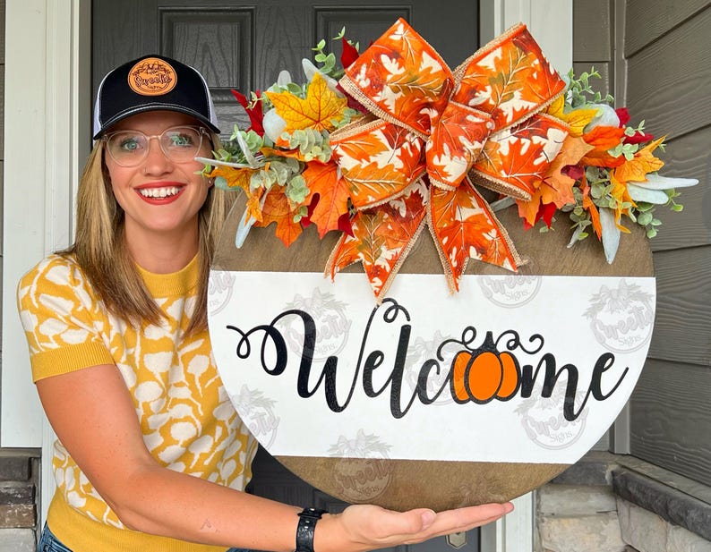 May include: A round wooden sign with a fall-themed wreath and the word 'Welcome' written in black cursive with a small orange pumpkin graphic. The wreath is made of orange and brown leaves, a white and orange bow, and green and white floral accents.