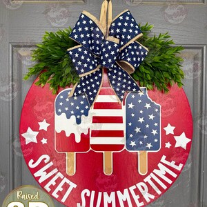 Op de afbeelding: Een rond rood houten deurbord met drie ijslolly-uitsparingen in Amerikaanse vlagpatronen. Het bord is bekroond met een grote blauw-witte strik met sterrenpatroon van jute en groen kunstblad. De tekst SWEET SUMMERTIME staat in het wit op het bord.
