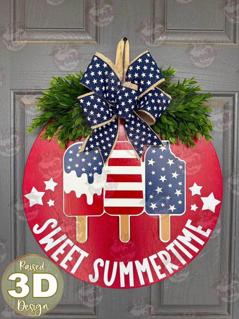 Op de afbeelding: Een rond rood houten deurbord met drie ijslolly-uitsparingen in Amerikaanse vlagpatronen. Het bord is bekroond met een grote blauw-witte strik met sterrenpatroon van jute en groen kunstblad. De tekst SWEET SUMMERTIME staat in het wit op het bord.