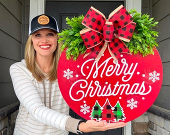 May include: A red round wooden door hanger with the text "Merry Christmas" in white cursive lettering. The door hanger is decorated with a red and black buffalo plaid bow, green foliage, and white snowflake accents.