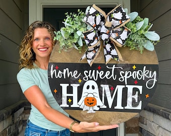 May include: A round wooden door sign featuring the text home sweet spooky HOME in white and script lettering. A white ghost illustration holding an orange pumpkin is centered. The sign is decorated with a black and white ghost patterned bow and green foliage.