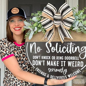 May include: A round wooden door sign with the text "No Soliciting" in white. The sign has a black background with a brown top section, a large bow, and greenery. The text also reads "Don't knock or ring doorbell, don't make it weird, seriously, friends & deliveries welcome."