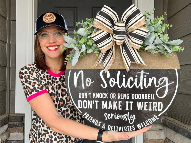 May include: A round wooden door sign with the text "No Soliciting" in white. The sign has a black background with a brown top section, a large bow, and greenery. The text also reads "Don't knock or ring doorbell, don't make it weird, seriously, friends & deliveries welcome."