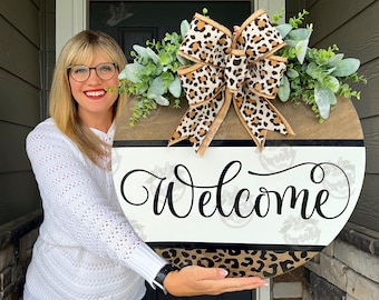 May include: Round wooden welcome sign with a leopard print bow and greenery. The sign has a brown top, a white center with the word "Welcome" in black script, and a leopard print bottom.