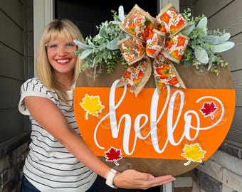 May include: A round wooden fall-themed sign with an orange half-circle and the word "hello" in white script. The sign is decorated with leaf accents and a decorative bow with fall colors.