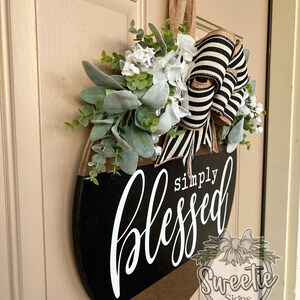 May include: A round wooden door sign with the words "simply blessed" in white script. The sign is black and brown with a floral arrangement and a black and white striped bow.