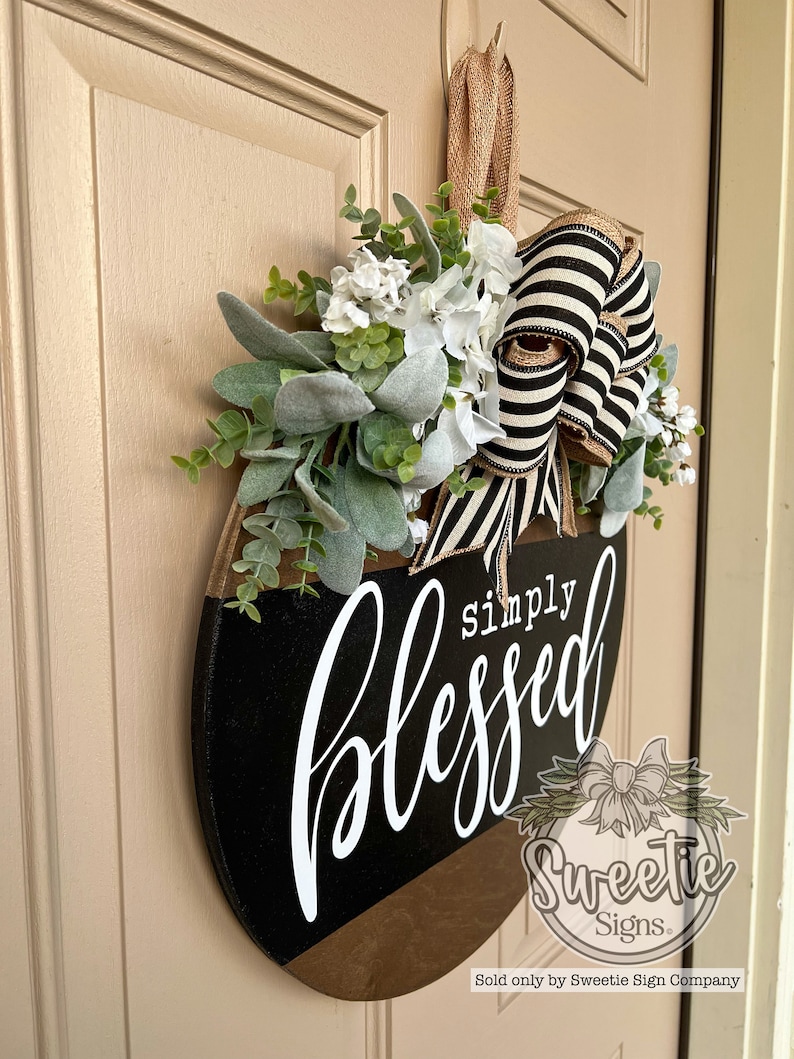 May include: A round wooden door sign with the words "simply blessed" in white script. The sign is black and brown with a floral arrangement and a black and white striped bow.