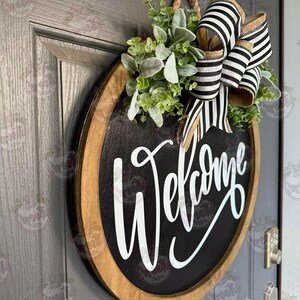 May include: A round wooden door sign with a black center and a natural wood frame. The word Welcome is written in white script lettering. The sign is decorated with a cluster of green faux leaves and a large bow made of black and white striped ribbon.
