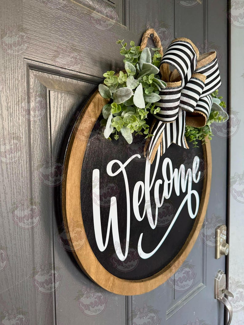 May include: A round wooden door sign with a black center and a natural wood frame. The word Welcome is written in white script lettering. The sign is decorated with a cluster of green faux leaves and a large bow made of black and white striped ribbon.