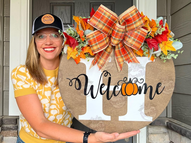 May include: A round wooden door hanger with a white letter H and the word 'Welcome' with a pumpkin in the middle of the word. The door hanger is decorated with a plaid bow and fall leaves.