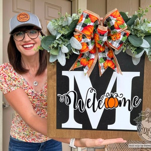 May include: A wooden welcome sign with a white letter "M" and the word "Welcome" with a pumpkin graphic. The sign is decorated with a burlap bow and a wreath of faux greenery with a black and white checkered ribbon.