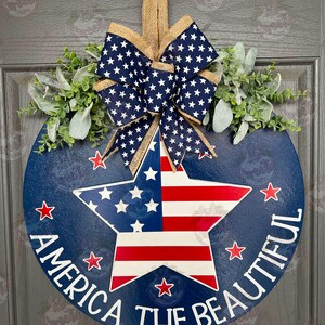 May include: A blue and red star with white stripes on a blue round wooden door hanger. The door hanger has the text "America the Beautiful" written around the star. The door hanger is decorated with a burlap bow with white stars on a navy blue background.