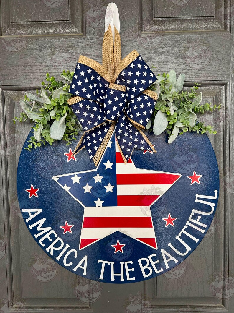 May include: A blue and red star with white stripes on a blue round wooden door hanger. The door hanger has the text "America the Beautiful" written around the star. The door hanger is decorated with a burlap bow with white stars on a navy blue background.