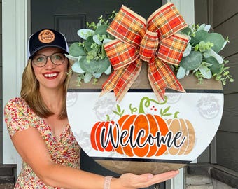 May include: A round wooden welcome sign with a pumpkin design. The sign features the word "Welcome" in a script font, with orange and tan pumpkins. It is decorated with a plaid bow and greenery.