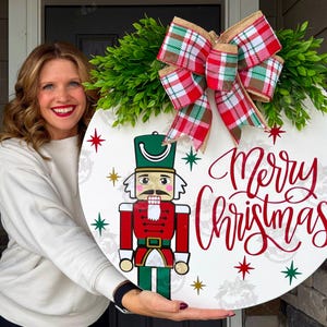 May include: A round, white Christmas door sign featuring a nutcracker illustration and the words "Merry Christmas" in red script. The sign is adorned with a green wreath, a plaid bow, and star accents.