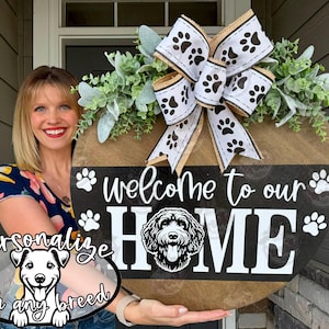 May include: A round wooden welcome sign with the text "Welcome to our HOME" and a dog illustration. The sign is decorated with a large bow with paw prints and greenery. The sign also has the text "Personalize with any breed".
