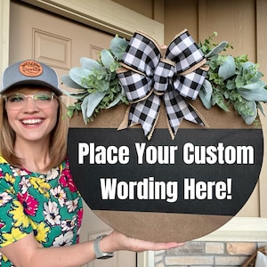 May include: Round wooden sign with a black and brown design, adorned with a buffalo check bow and greenery. The sign reads "Place Your Custom Wording Here!" in white text, perfect for personalized home decor.