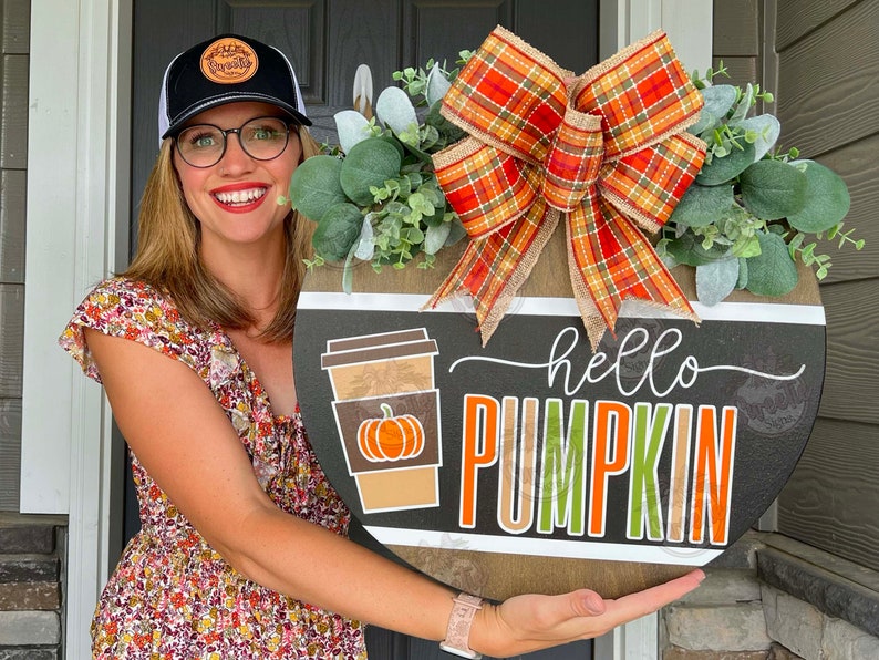 May include: A round wooden sign with a black background and white lettering. The sign says 'hello pumpkin' and has a coffee cup with a pumpkin inside. The sign is decorated with a plaid bow and green leaves.