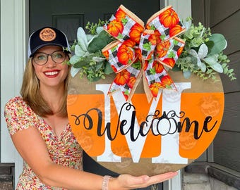 May include: A round wooden welcome sign with an orange and brown pumpkin theme. The sign features a large white "M" and the word "Welcome" in black script. A decorative bow with pumpkin and plaid patterns and greenery adorns the top.