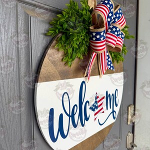 May include: A round wooden door sign featuring a white lower section with the blue script text Welcome, where the letter o is replaced by a star with a blue and white field and red stripes. A large bow with American flag patterns sits above.