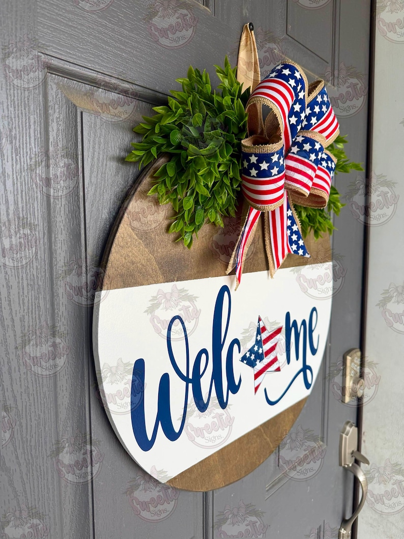 May include: A round wooden door sign featuring a white lower section with the blue script text Welcome, where the letter o is replaced by a star with a blue and white field and red stripes. A large bow with American flag patterns sits above.