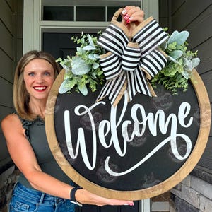 May include: A round wooden door sign with a black center featuring the white script text Welcome. The sign is decorated with a large black and white striped fabric bow and artificial green eucalyptus leaves attached to the top of the frame.