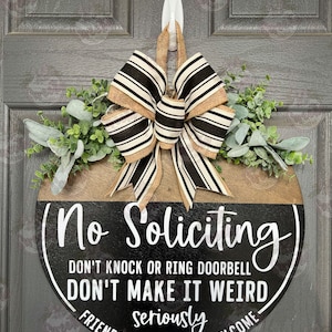 May include: A round wooden door sign with the text "No Soliciting" in white lettering. The sign has a black and brown color scheme, a decorative bow, and greenery. The sign also includes the phrases "Don't Knock or Ring Doorbell", "Don't Make It Weird", and "Friends & Deliveries Welcome".