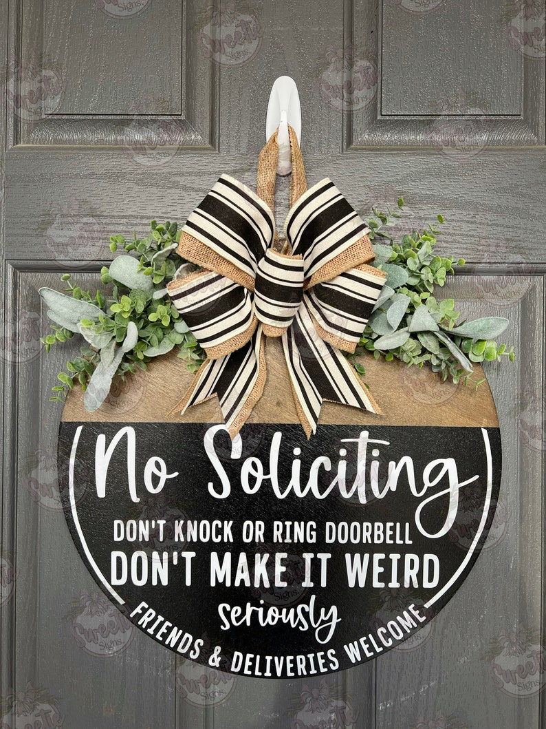 May include: A round wooden door sign with the text "No Soliciting" in white lettering. The sign has a black and brown color scheme, a decorative bow, and greenery. The sign also includes the phrases "Don't Knock or Ring Doorbell", "Don't Make It Weird", and "Friends & Deliveries Welcome".