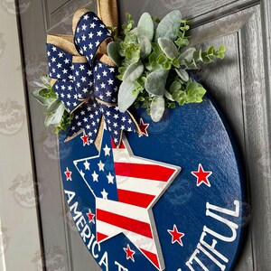 May include: A blue and red star with white stripes, surrounded by white stars on a blue background. The text "America the Beautiful" is written around the star. A blue and white star-patterned bow with burlap accents and green foliage are attached to the top of the round wooden sign.