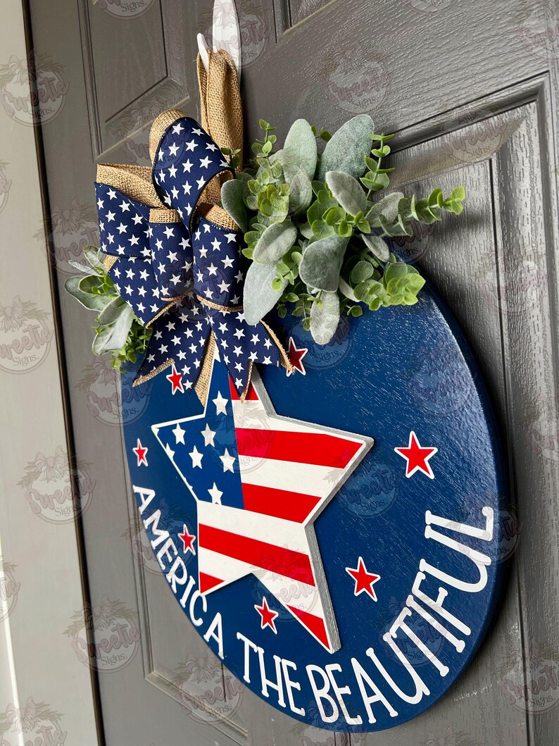 May include: A blue and red star with white stripes, surrounded by white stars on a blue background. The text "America the Beautiful" is written around the star. A blue and white star-patterned bow with burlap accents and green foliage are attached to the top of the round wooden sign.