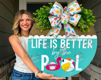 Pode incluir: Uma placa redonda de madeira com uma metade inferior turquesa e uma metade superior castanha. A seção turquesa diz "LIFE IS BETTER by the POOL" com um flamingo e um gráfico de bola de praia. A placa é decorada com uma coroa verde e um laço com desenhos de picolés.
