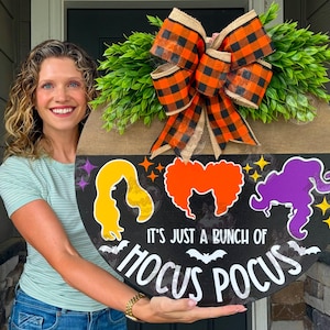May include: A round wooden door sign featuring a black background with three colorful hair silhouettes in yellow, orange, and purple. The text reads IT'S JUST A BUNCH OF HOCUS POCUS in white, accented by small bats and stars, topped with a large orange and black plaid bow and green foliage.