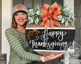 May include: A round wooden door sign featuring a black horizontal centre stripe with the white text Happy Thanksgiving and an orange pumpkin graphic. The sign is decorated with a large orange and brown plaid bow and green foliage.