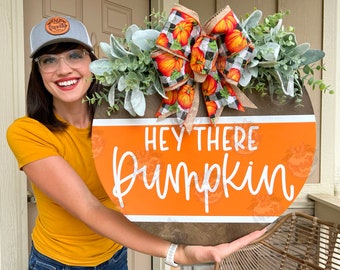 May include: A round wooden door hanger with an orange background and white text that reads "Hey there Pumpkin". The door hanger is decorated with a large orange and black plaid bow with a burlap ribbon and a green leafy garland.