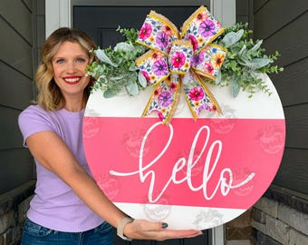 May include: A round wooden door sign featuring a white and hot pink horizontal striped background with the word hello written in white cursive script. The sign is decorated with a large floral patterned bow and green faux eucalyptus foliage.