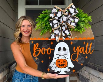 May include: A round wooden door sign featuring a black background with the orange text Boo y'all and a white ghost holding an orange jack-o'-lantern. The sign is topped with a large bow patterned with white ghosts and green faux greenery.