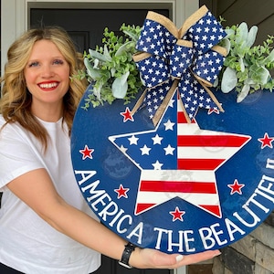 May include: A blue and red star-shaped door hanger with a white background. The star has red and white stripes and white stars. The text "America the Beautiful" is written in white around the star. The door hanger is decorated with a blue and white bow and greenery.